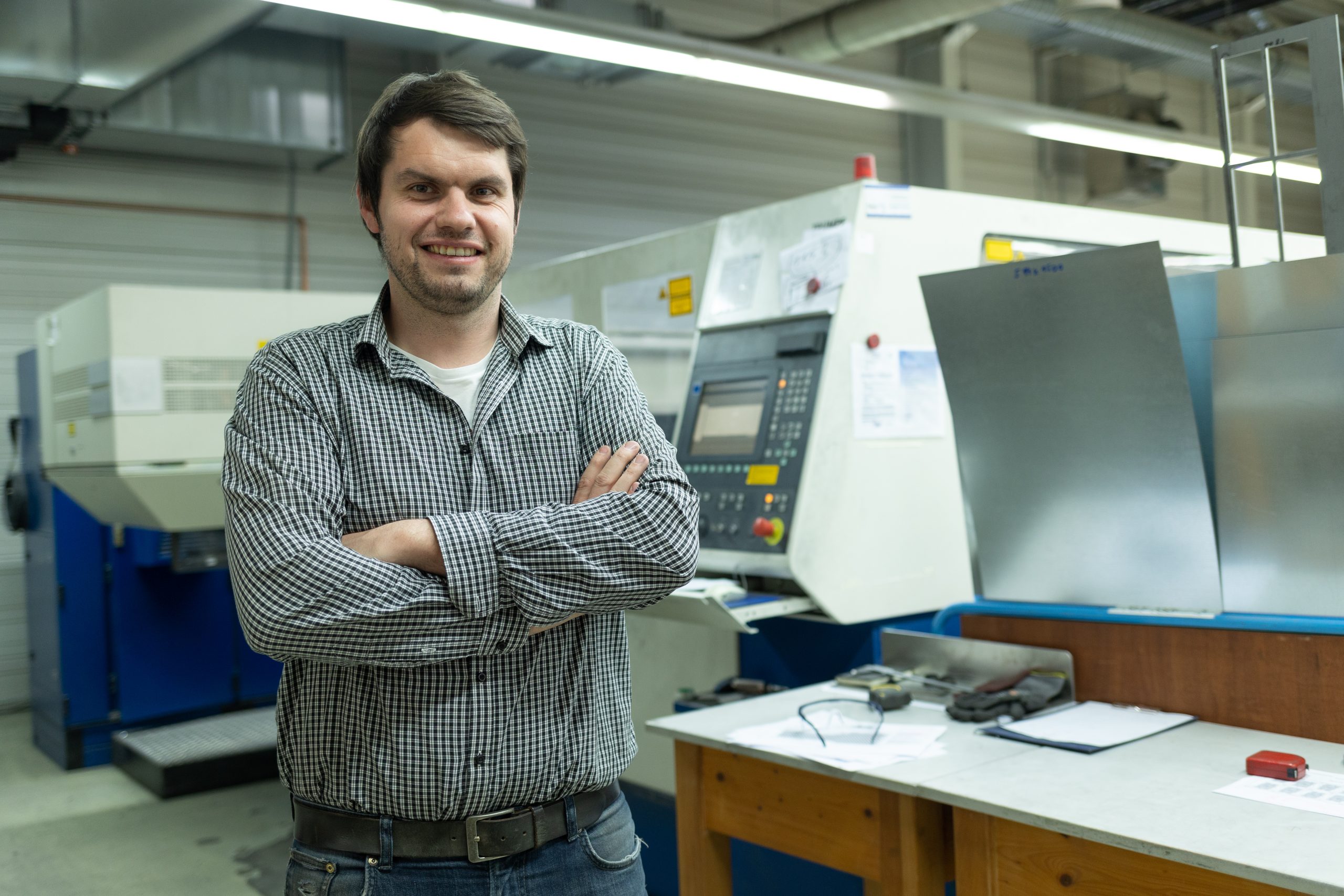Zufriedener Mitarbeiter steht in einer Produktionshalle mit CNC Maschinen. Er zeigt Mitarbeiterloyalität